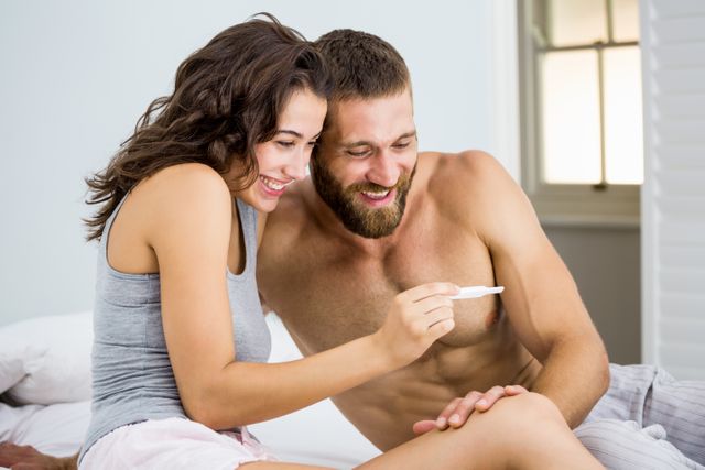 Happy couple looking at pregnancy test on bed at bedroom