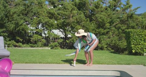 Woman placing sunscreen by pool edge wearing straw hat and striped cover-up, poolside care