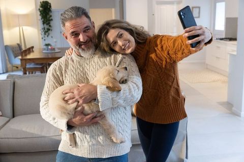 Cozy couple taking smartphone selfie while holding golden retriever puppy in modern home