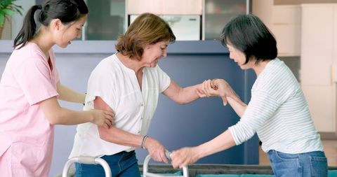 Senior woman using walker assisted by caregiver and friend indoors