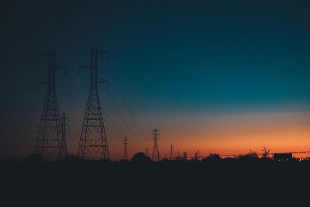 Transmission towers are silhouetted against a colorful sunset sky, creating a stark contrast between nature and modern technology. Ideal for illustrating themes of energy, power infrastructure, and rural landscapes. Suitable for use in energy industry materials, environmental discussions, and promotional materials for sustainable energy.