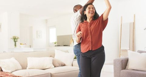 Joyful couple dancing in bright modern living room, woman spinning in rust blouse