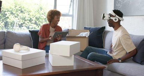 Smiling couple unpacking VR headset and tech boxes, laughing together on living room sofa