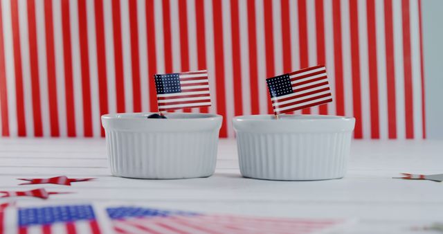 Decorative scene featuring two small American flags placed in white ramekins with a red and white striped background. Suitable for use in patriotic content related to US holidays such as Independence Day, July 4th celebrations, and other American-themed events. This can be used for social media posts, blog content, and advertising material to evoke a sense of national pride.