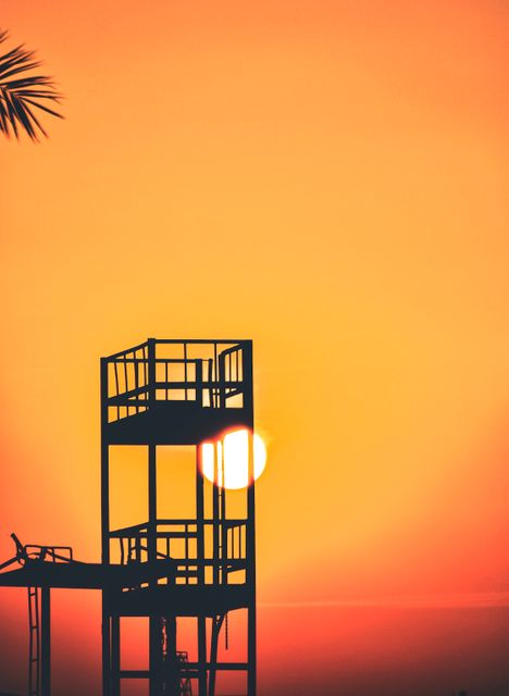 Lifeguard tower silhouette contrasts against vibrant orange sunset sky. Use for beach safety campaigns, vacation promotions, and serene evening scenes.