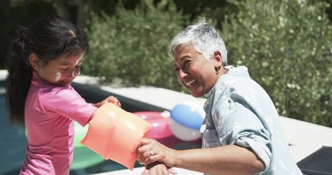 Grandparent helping child adjust orange arm float at backyard pool on sunny day