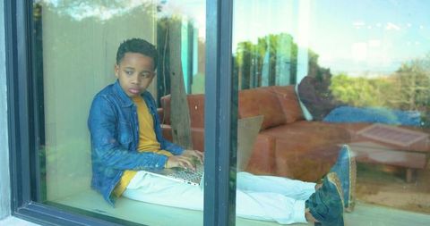 African american boy sitting on window seat using laptop while father resting on sofa