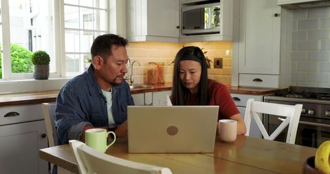 Asian couple working together on laptop at cozy kitchen table planning finances and collaborating