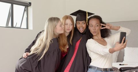 Graduating friends celebrating and taking selfie on bed wearing gowns and mortarboard