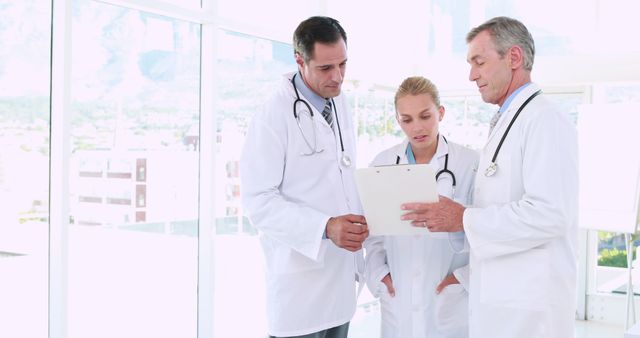 Two middle-aged Caucasian men and a young Caucasian woman, all dressed in medical attire, are discussing a document, with copy space. Their focused expressions and professional attire suggest they are healthcare professionals, doctors, engaged in a serious medical consultation.