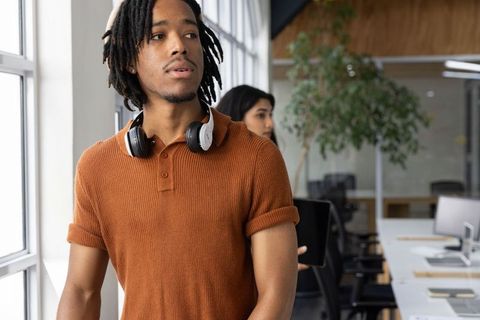 Young black professional standing by window wearing headphones holding tablet in office