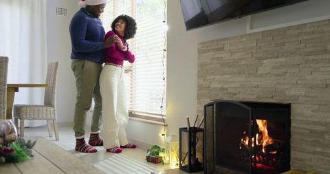 Diverse couple sharing cozy holiday moment by fireplace and window with santa hat