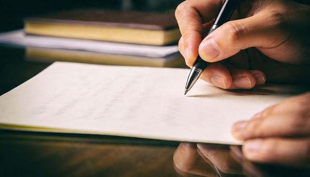 Right hand writing on paper with black ballpoint pen over reflective wooden desk close-up