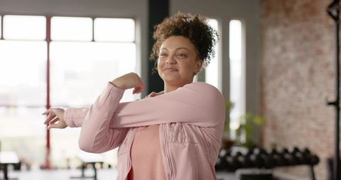 African american woman stretching arm across chest in gym wearing pink active jacket