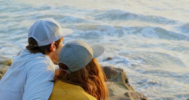 Couple enjoying a tranquil moment by the ocean waves, wearing casual hats. Ideal for themes of romance, relaxation, travel, and companionship. Can be used in travel brochures, wellness and lifestyle blogs, or advertisements promoting beach vacations and outdoor activities.
