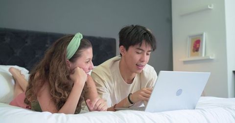 Young couple lounging on bed using laptop for streaming, planning, learning together
