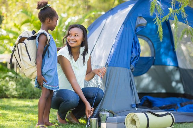 Mother and daughter enjoying a camping trip in a park. The mother is setting up a tent while the daughter, wearing a backpack, looks on happily. This image is perfect for promoting family bonding activities, outdoor adventures, camping gear, and summer vacations.