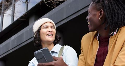 Smiling diverse couple sharing smartphone on urban sidewalk wearing mustard jacket and denim beanie