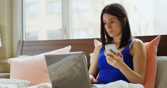 Woman wearing casual clothes, sitting on bed with pillows, multitasking using laptop and smartphone, working remotely from home. Great for illustrating remote work, technology use, comfort, or home office themes.