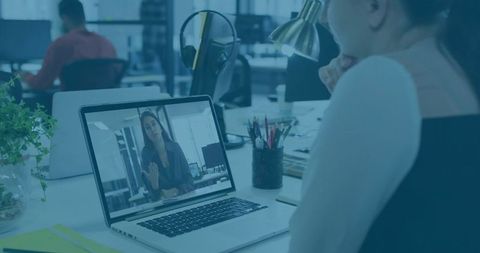 Woman joining remote video call in open-plan office, working at laptop with pens