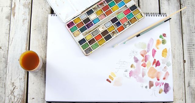 A set of watercolors and a paintbrush lie next to a sketchbook with color swatches and doodles on a wooden surface. Artistic tools like these inspire creativity and are essential for artists to experiment with color and technique.