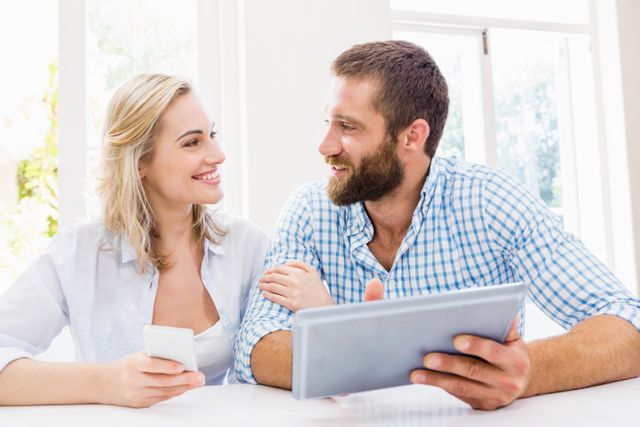 Couple sitting at home, smiling while using a digital tablet and smartphone. Ideal for use in articles or advertisements about modern technology, communication, relationships, and home lifestyle.