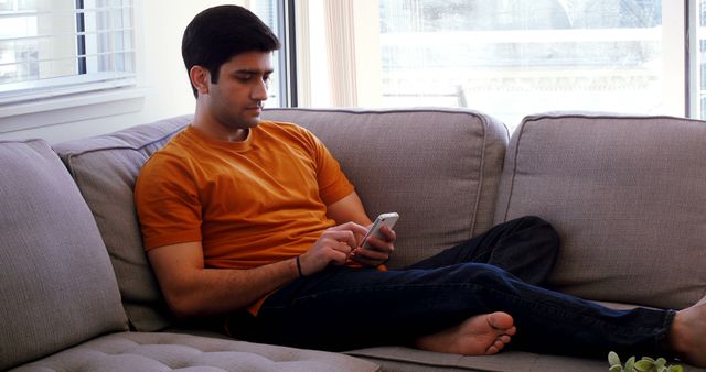 Young man sitting comfortably on couch while using smartphone. Ideal for themes of modern lifestyle, home comfort, technology usage, and relaxation.