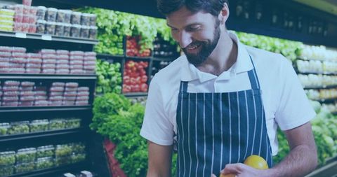 Smiling grocery clerk checking yellow bell pepper while stocking fresh produce in colorful aisle