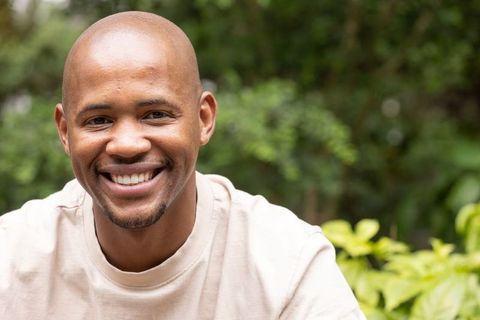 Smiling african american man in casual beige t-shirt outdoors with greenery and bokeh
