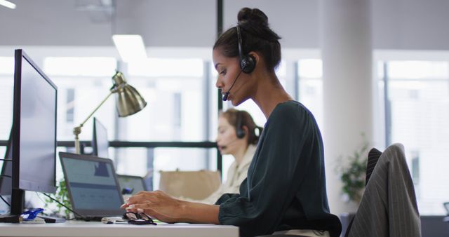 Biracial businesswoman sitting at desk talking using phone headset and computer in busy office - Download Free Stock Photos Pikwizard.com