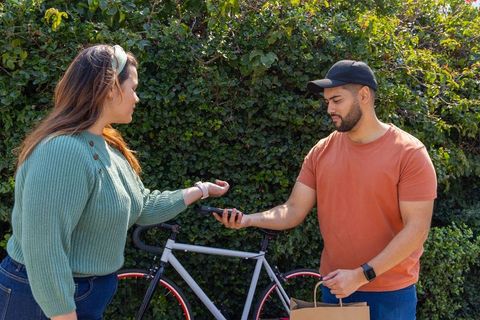 Adults exchanging smartphone and grocery bag beside bike on sunny sidewalk with greenery