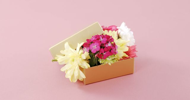 A colorful assortment of fresh flowers is neatly arranged in a small open gift box on a pink background, with copy space. Flowers like these are often given as a token of appreciation or to celebrate special occasions.