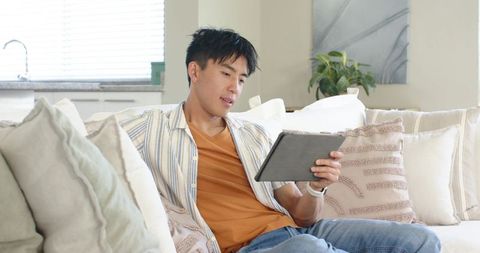 Young asian man relaxing on sofa using tablet for streaming, reading, and remote work