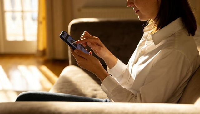 Woman using smartphone on sofa in sunlit living room cozy home casual morning