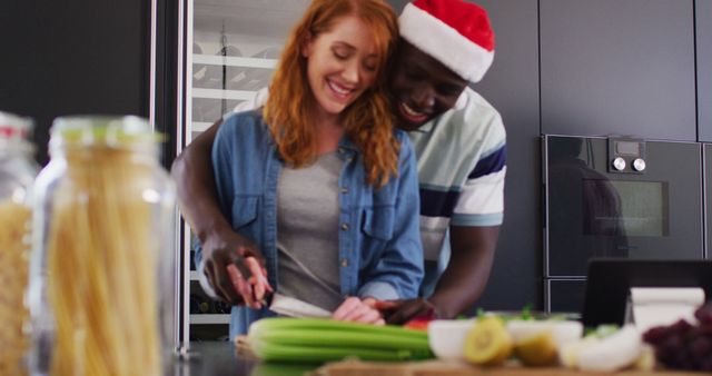 Interracial couple cooking in a contemporary kitchen, exuding love and happiness during the holiday season. Woman is cutting vegetables while man playfully hugs her, wearing a Santa hat. Perfect for depicting themes of togetherness, holiday celebrations, love, and multicultural relationships in advertising, blog posts, and social media.