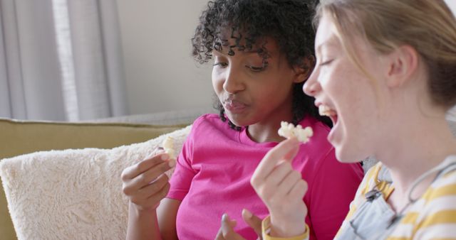 Two friends sitting on a cozy sofa are enjoying popcorn together. They appear comfortable and happy, embodying a moment of leisure and joy. Use this image to promote concepts of friendship, bonding, relaxation, and enjoying simple pleasures. Perfect for advertisements for snacks, living room furniture, or lifestyle blogs.