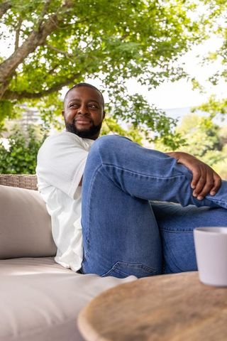 African american man relaxing on outdoor lounge sofa with mug under leafy patio