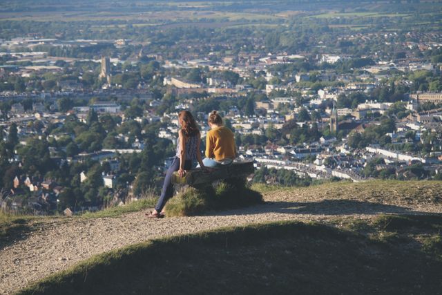 Two women enjoying peaceful view, sitting on bench at mountain peak. Ideal for themes of travel, sightseeing, leisure, relaxation, and nature appreciation. Useful in promotional material for outdoor activities, lifestyle blogs, and tourism campaigns.