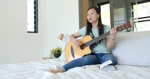 Asian woman composing and playing acoustic guitar on bed in bright minimalist bedroom