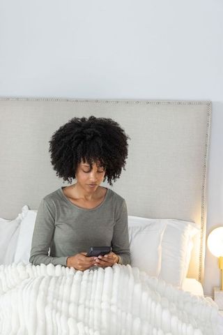 African american woman sitting in bed holding smartphone under cozy blanket in modern bedroom