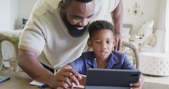 A man assists his young boy with online learning on a tablet. This can be used for images relating to family time, online education, homeschooling, remote learning, father-son relationship, and parental support in education.