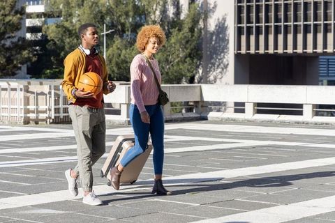 African american friends walking with basketball and rolling suitcase across urban plaza