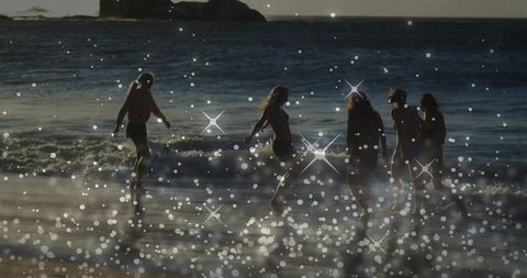 Backlit friends wading on sunlit shoreline at golden hour with sparkling bokeh reflections