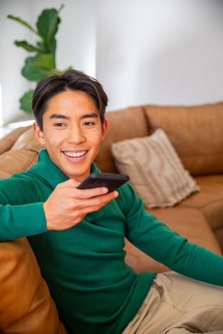Smiling asian man relaxing on tan sofa holding tv remote in cozy modern living room