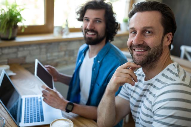 Two smiling men using digital tablet and laptop at coffee shop