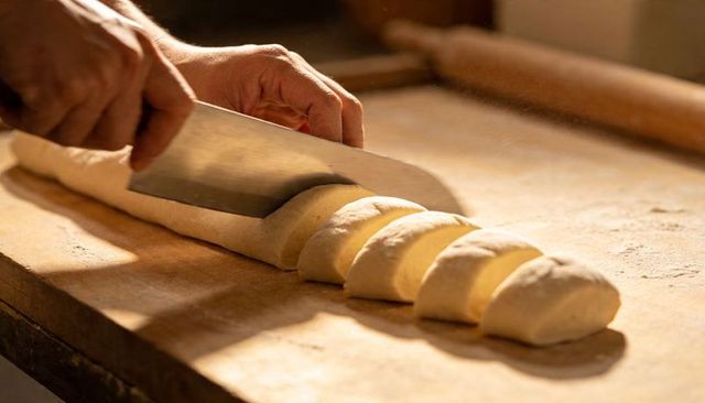 Baker slicing dough log with cleaver on floured wooden board rustic bakery close-up