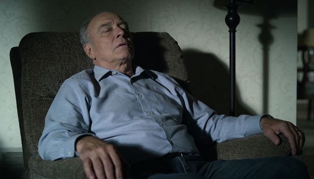 Dozing senior man reclining in armchair at home with lamp light and moody shadows
