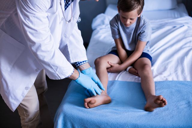 Male doctor wearing gloves examining young boy's leg in a hospital room. The boy is sitting on a bed, looking concerned. This image can be used for healthcare, medical, pediatric care, and hospital-related content. It is suitable for illustrating articles about child health, medical examinations, and professional healthcare services.