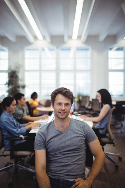 Portrait of man smiling at camera while colleagues working in background at the office