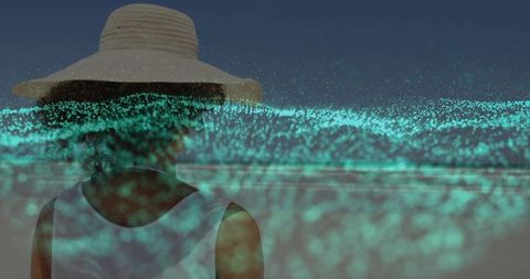 Woman in straw hat overlaid with glowing turquoise particles at dusk beach